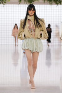 Model walking a fashion show in a beige oversized jacket, pink lace top, and pale green bubble shorts with strappy nude sandals.