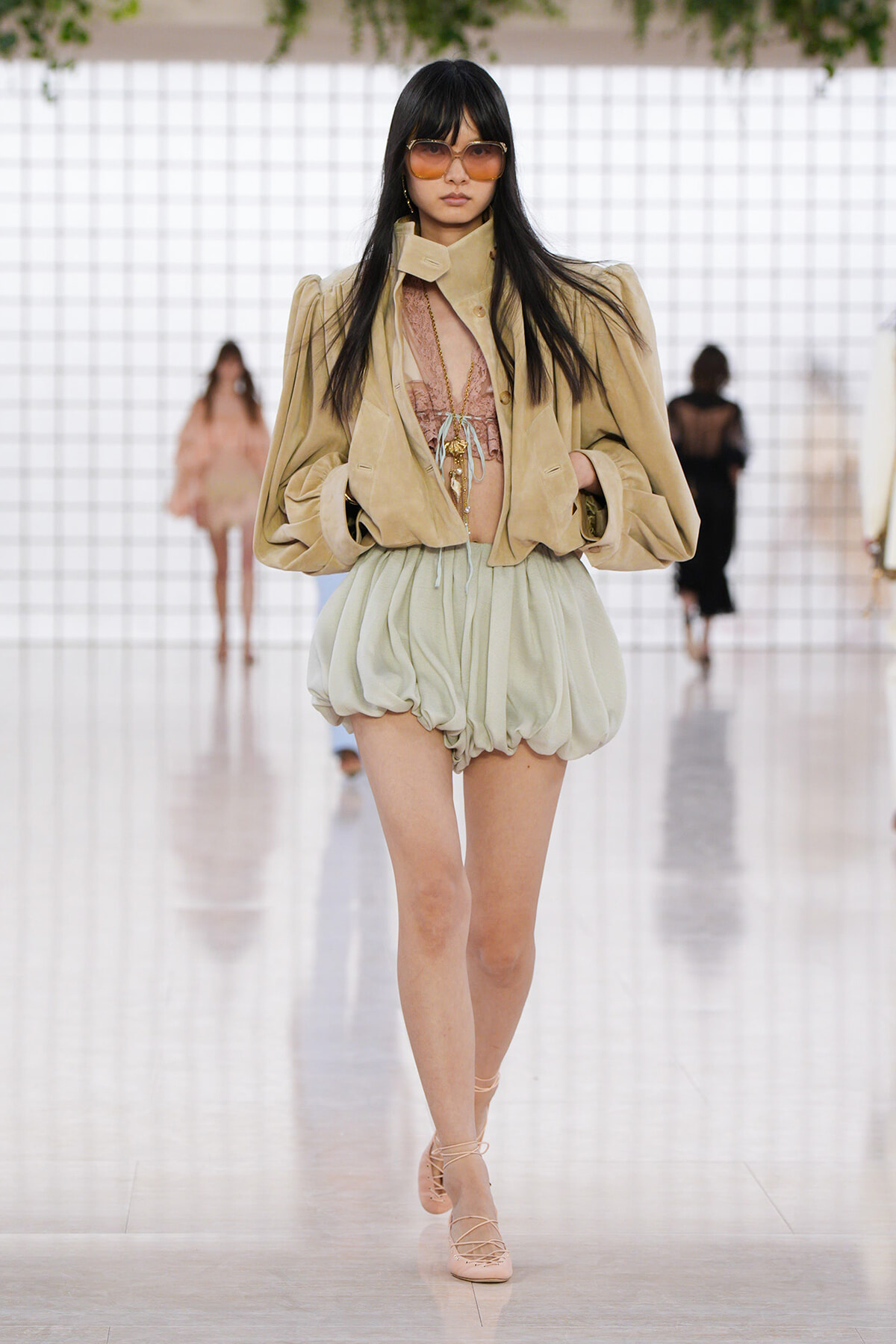 Model walking a fashion show in a beige oversized jacket, pink lace top, and pale green bubble shorts with strappy nude sandals.