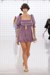 Model walking runway in a lilac off-the-shoulder mini dress with puff sleeves, cinched waist, and ruffled hem, accessorized with a small straw bag and tan sandals