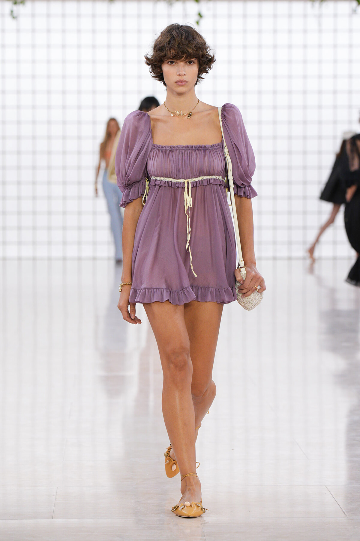 Model walking runway in a lilac off-the-shoulder mini dress with puff sleeves, cinched waist, and ruffled hem, accessorized with a small straw bag and tan sandals