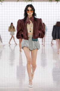 Model walking a runway in a burgundy leather jacket, cream lace top, and voluminous gray shorts, paired with white strappy heels and oversized sunglasses.