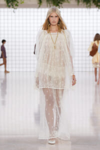 Model walking a fashion runway in a long, sheer white lace dress with floral embroidery, gold jewelry, and white shoes.