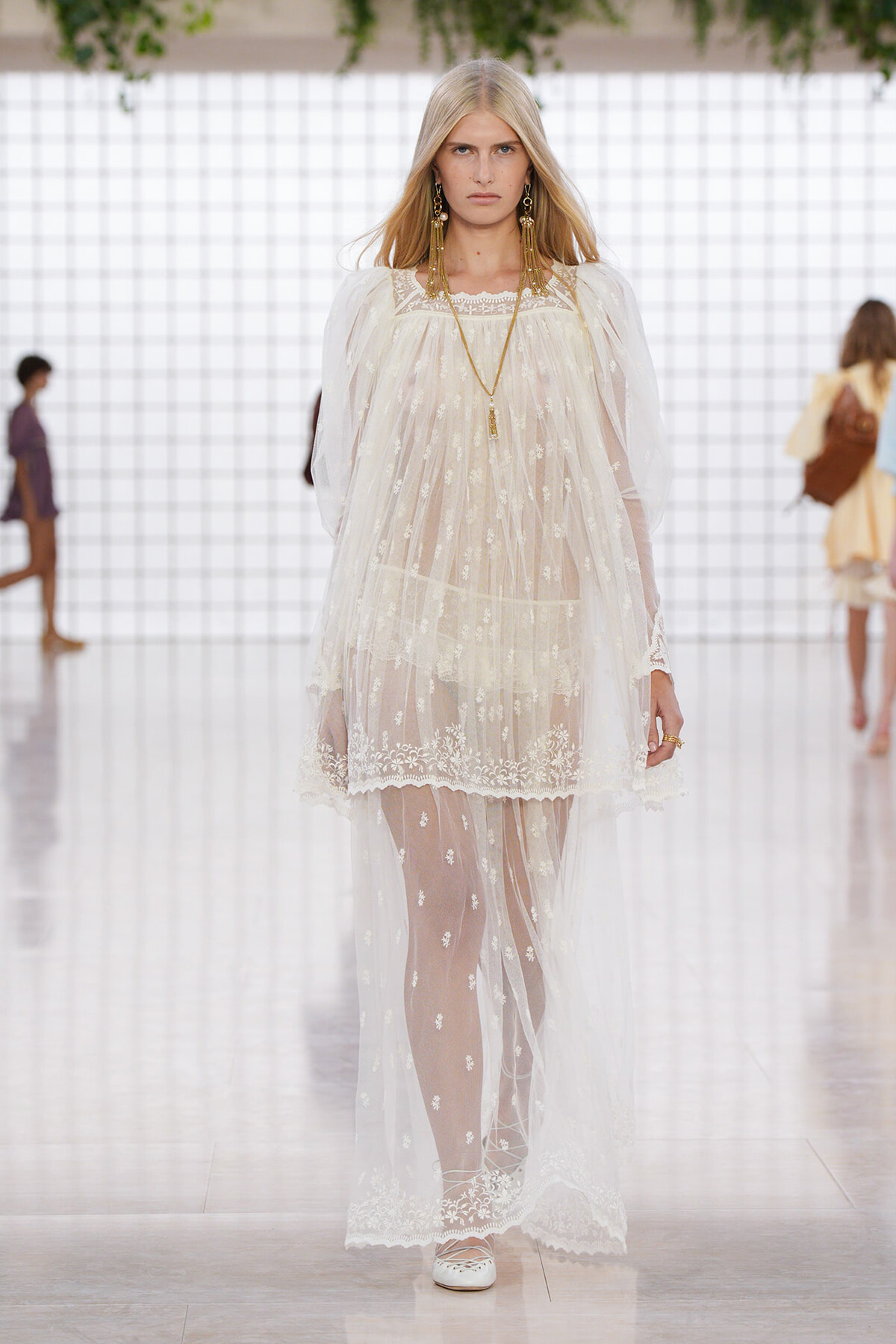 Model walking a fashion runway in a long, sheer white lace dress with floral embroidery, gold jewelry, and white shoes.