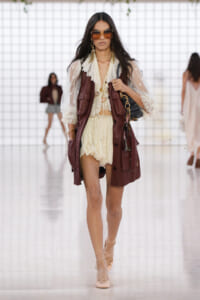 Model walking a runway in a cream lace blouse and matching shorts, layered with a burgundy leather vest, gold jewelry, oversized sunglasses, and nude lace-up heels, carrying a black bag.