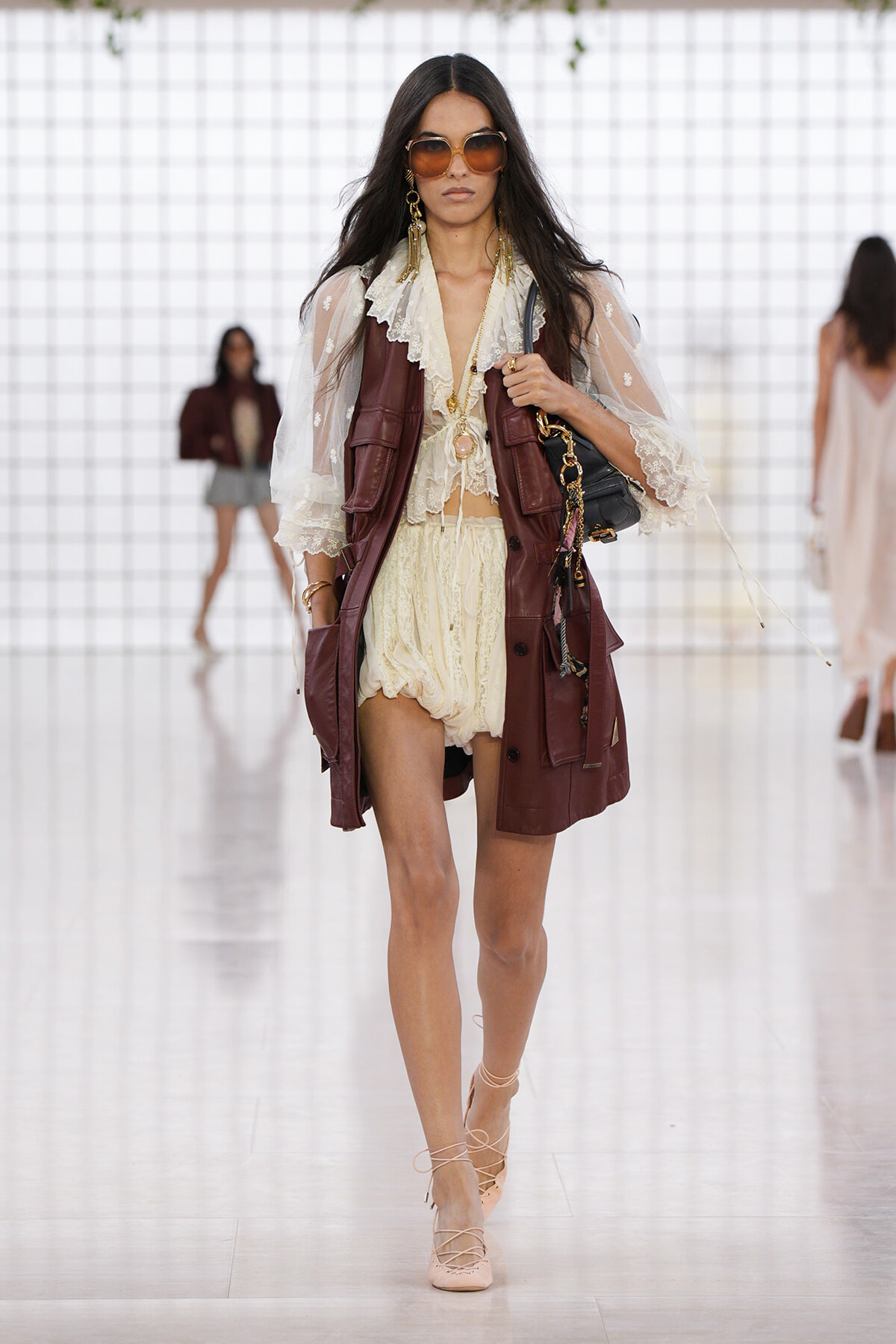 Model walking a runway in a cream lace blouse and matching shorts, layered with a burgundy leather vest, gold jewelry, oversized sunglasses, and nude lace-up heels, carrying a black bag.