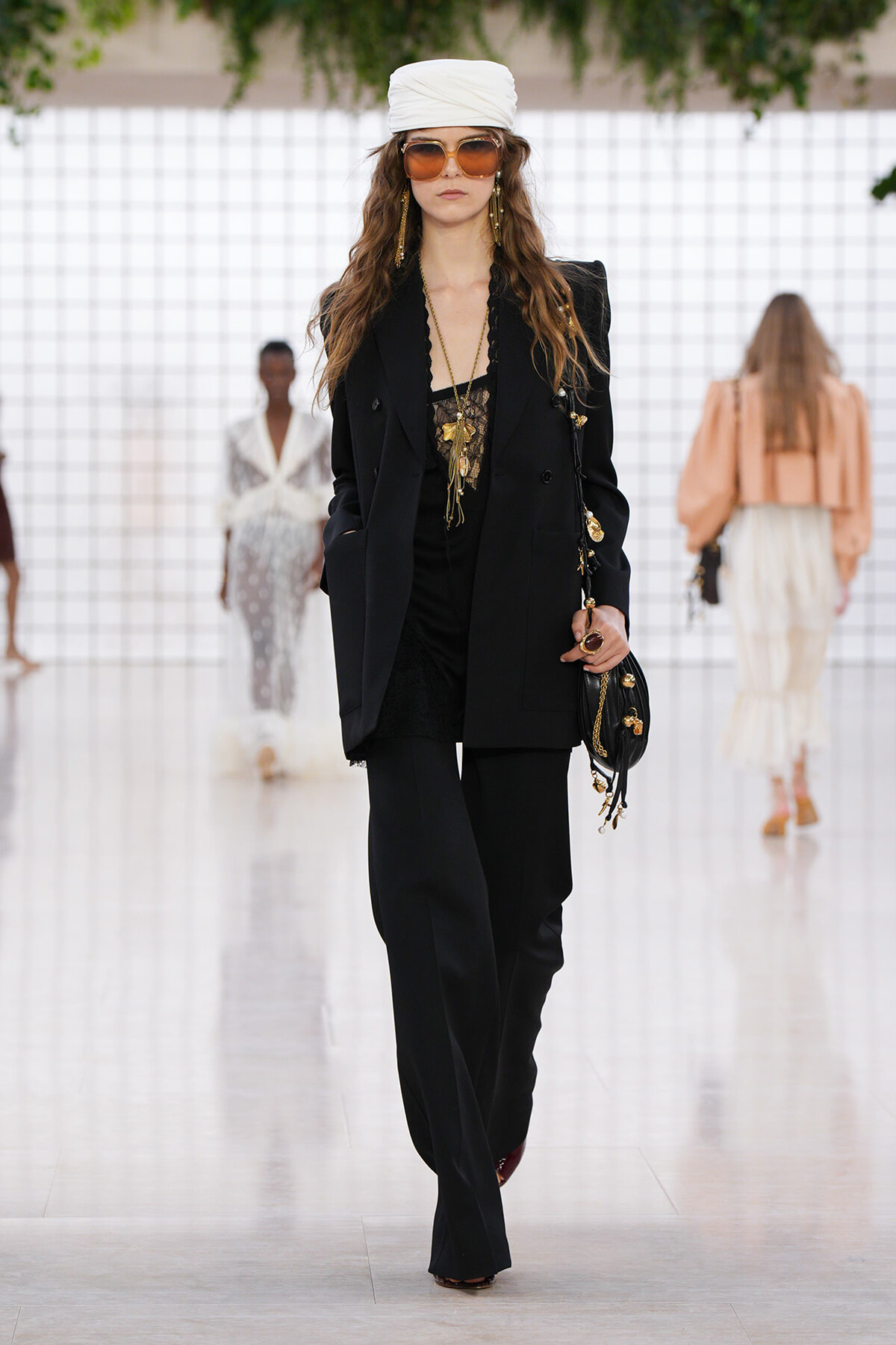 Female runway model wearing a black pantsuit with a long blazer, lace top, and gold jewelry, paired with a white head wrap and oversized sunglasses while carrying a black handbag.