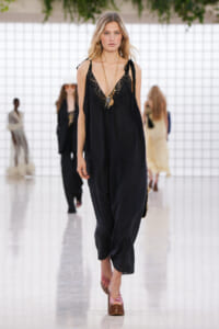 Model walking a runway in a loose black lace-trim jumpsuit with layered gold necklaces and pink platform sandals