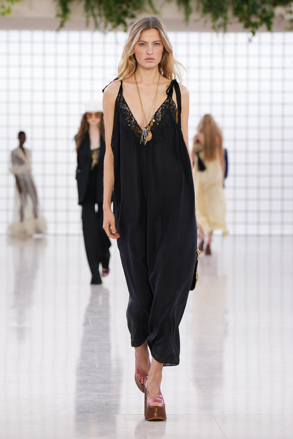 Model walking a runway in a loose black lace-trim jumpsuit with layered gold necklaces and pink platform sandals