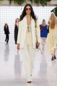 Model walking the runway in a cream pantsuit with a lace top and oversized sunglasses with gold accessories.