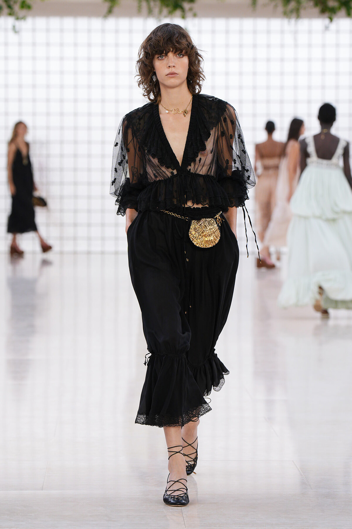 Model walking a runway in sheer black lace blouse with puff sleeves and wide gathered black pants, accessorized with a gold belt bag and lace-up heels.