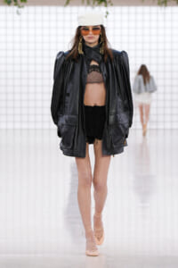 Runway model in an oversized black leather jacket over a lace bralette and black shorts, with a white turban and large sunglasses.