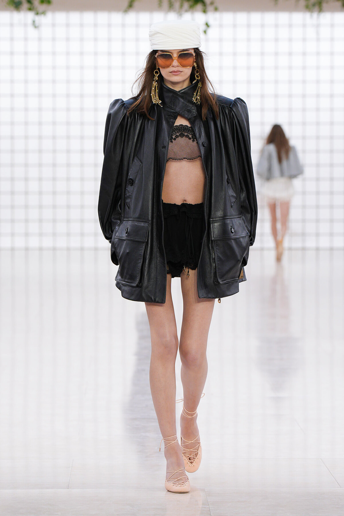 Runway model in an oversized black leather jacket over a lace bralette and black shorts, with a white turban and large sunglasses.