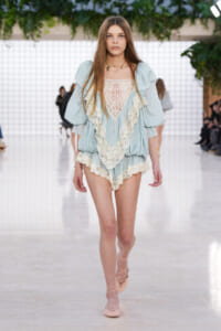 Model walks a fashion runway wearing a pale blue lace-trim romper with puff sleeves and beige lace-up flats.