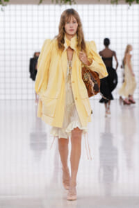 Model walking runway in a pale yellow oversized coat over a cream lace dress, carrying a brown handbag with charms.