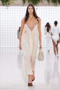 Model walks the runway in a cream satin jumpsuit with pink lace neckline, holding a woven beige handbag.