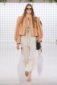 Model walking a runway in a peach oversized jacket over a cream lace top and gathered cream pants, carrying a black crossbody bag and wearing oversized sunglasses.