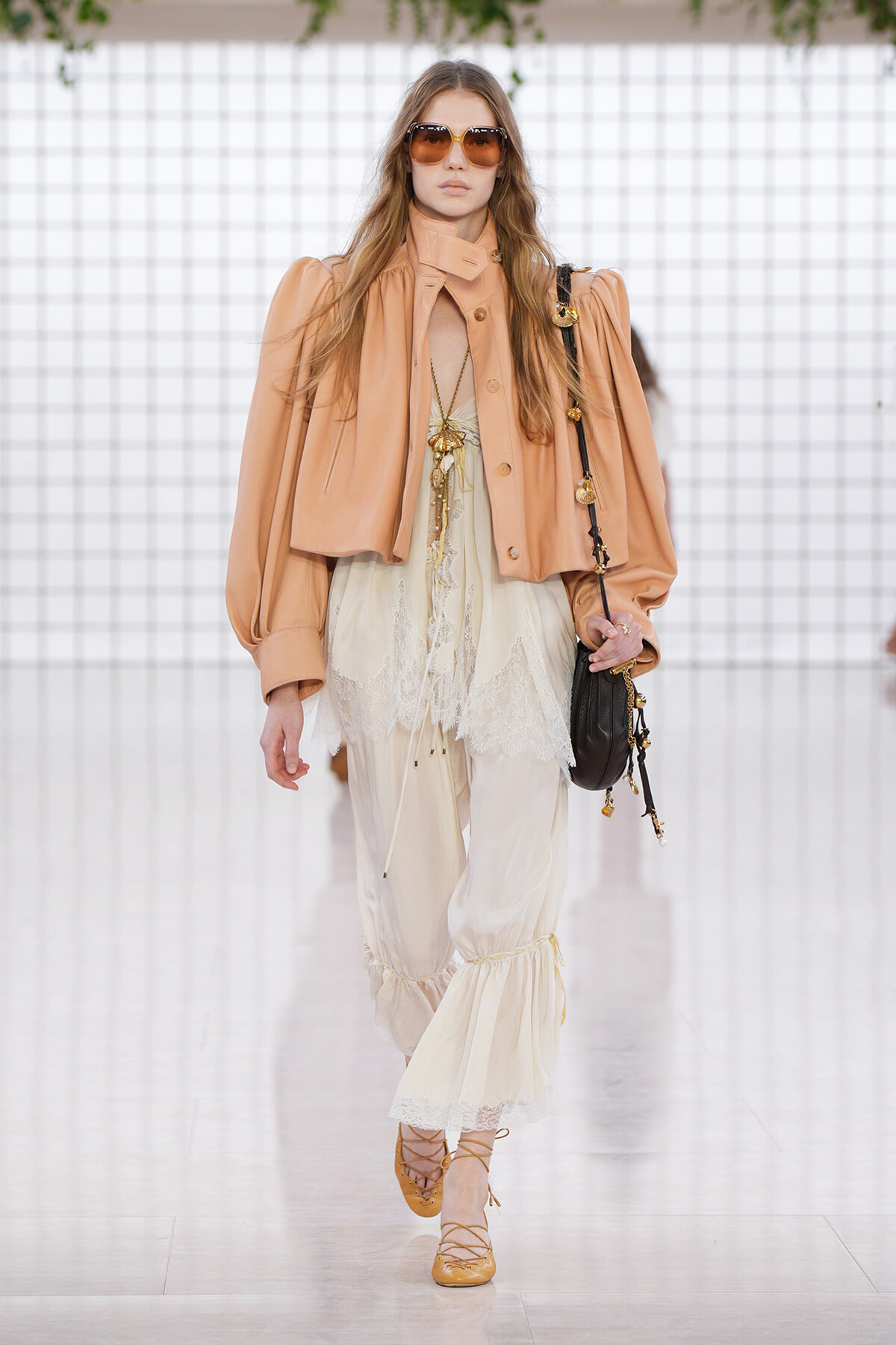 Model walking a runway in a peach oversized jacket over a cream lace top and gathered cream pants, carrying a black crossbody bag and wearing oversized sunglasses.