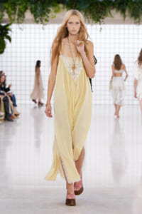 Model on a runway wearing a flowing pale yellow dress with lace bodice, gold jewelry, and pink platform sandals.