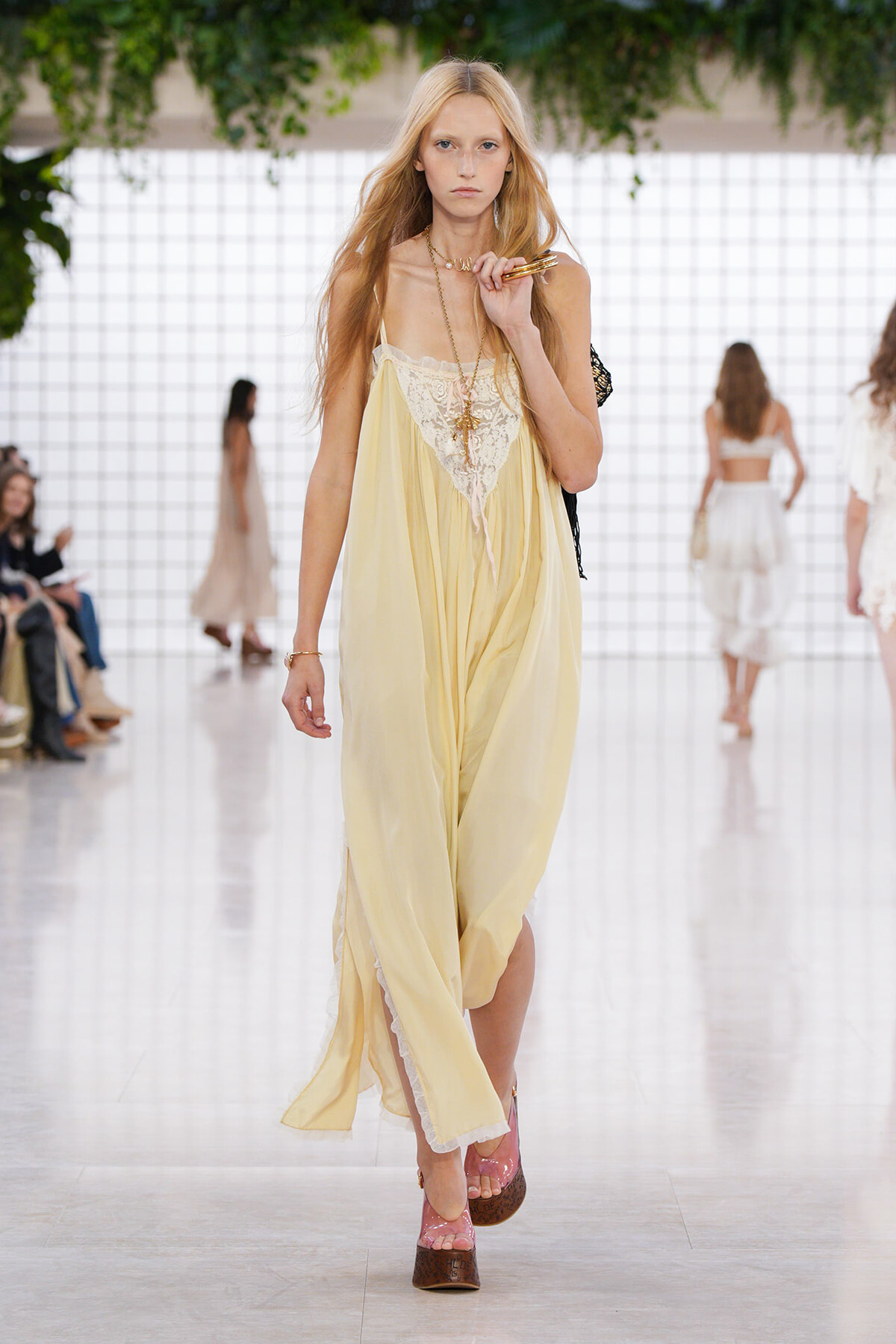 Model on a runway wearing a flowing pale yellow dress with lace bodice, gold jewelry, and pink platform sandals.