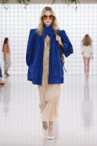 Model on a runway wearing a bright blue oversized coat over a beige layered dress, with large sunglasses and white lace-up shoes.