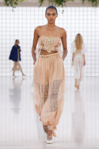 Model walking a fashion runway in a peach lace bustier and sheer lace-panel pants with tied ankles and white lace-up heels.