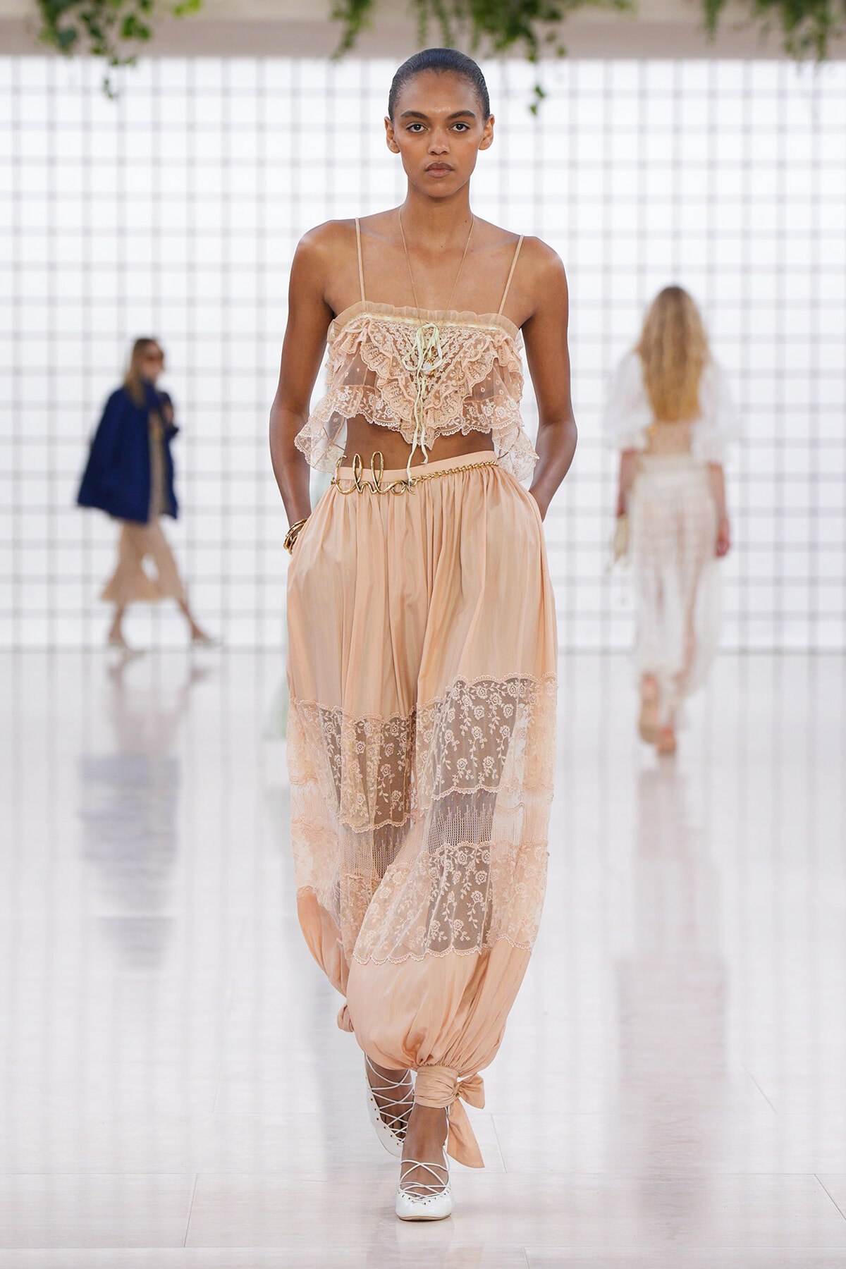 Model walking a fashion runway in a peach lace bustier and sheer lace-panel pants with tied ankles and white lace-up heels.