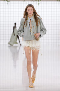Model walking a runway in a pale gray puff-sleeve jacket, ivory lace shorts, and tan lace-up sandals. 450px view on a white grid backdrop.