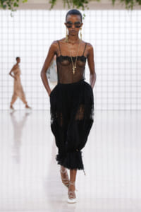 Model walking a runway in a sheer black lace top and voluminous lace-trim skirt, white lace-up heels, and gold jewelry with oversized sunglasses.
