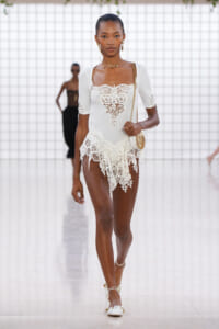 Model walking runway in a white lace dress with scalloped hem, paired with gold jewelry and a small gold chain purse; bright, minimalist backdrop.