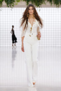 Female model walking a runway in an all-white lace blouse with puff sleeves and high-waisted white pants, accessorized with gold jewelry.