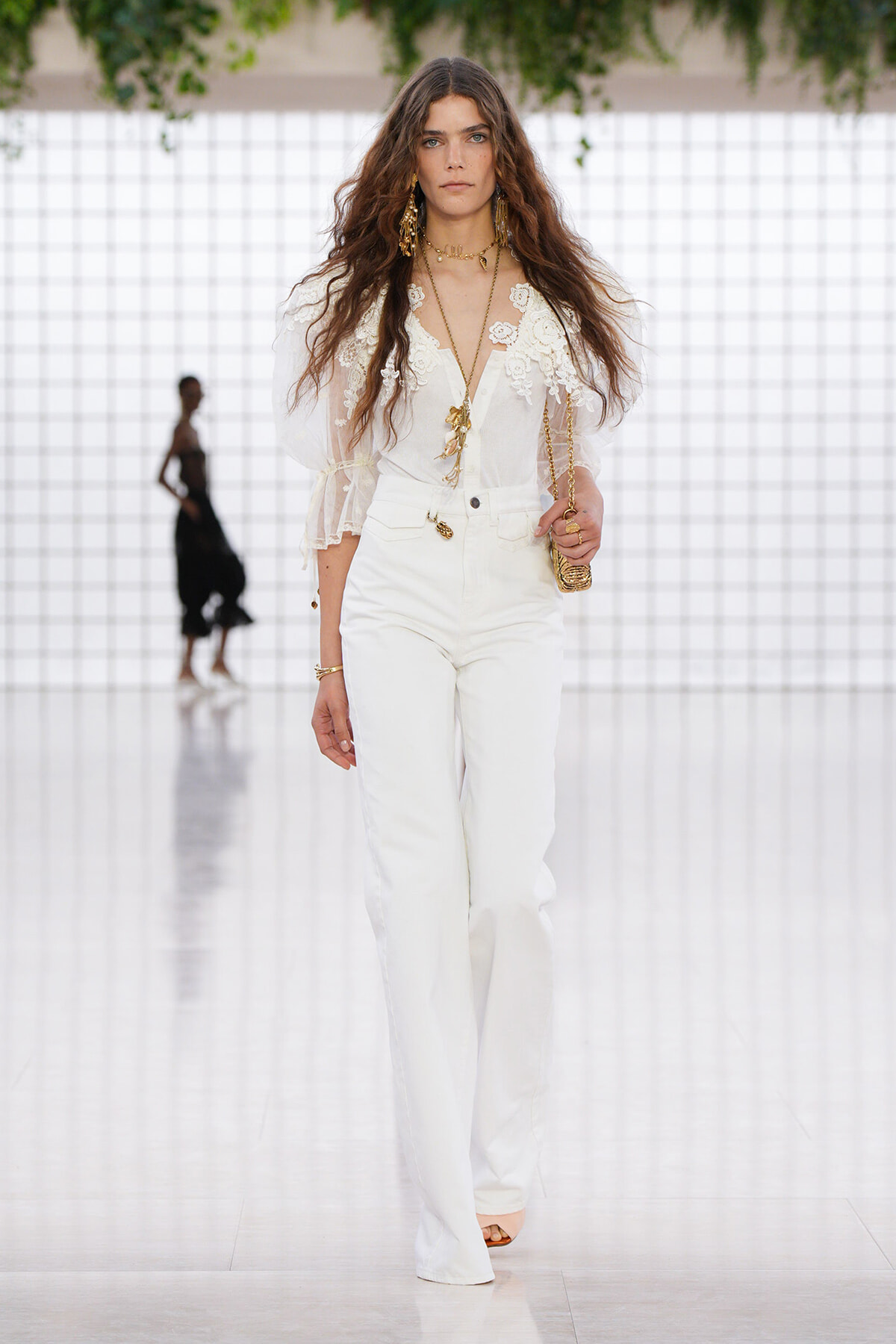 Female model walking a runway in an all-white lace blouse with puff sleeves and high-waisted white pants, accessorized with gold jewelry.