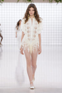Model walking a runway in a white lace dress with floral appliqués and sheer panels on a fashion show backdrop.