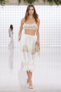 Model walks a runway in a white lace two-piece set: sheer lace crop top and flowing skirt with lace panels, beige sandals and a small bag.