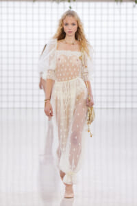 Model walks the runway in a sheer ivory lace dress with dotted embroidery and puffy sleeves, paired with gold jewelry and a small handbag.