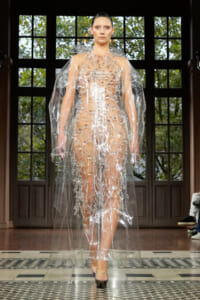 Model walks a runway wearing a transparent PVC gown with a bead-studded bodice and matching clear cape, in a dark-paneled venue with a checkered floor.