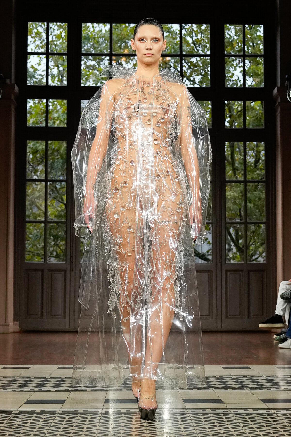 Model walks a runway wearing a transparent PVC gown with a bead-studded bodice and matching clear cape, in a dark-paneled venue with a checkered floor.