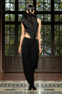 Model walking in a black, asymmetrical backless top and wide-leg pants on a runway with large paneled doors behind.