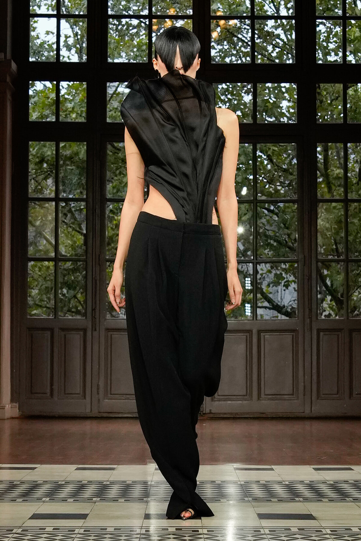 Model walking in a black, asymmetrical backless top and wide-leg pants on a runway with large paneled doors behind.