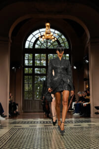 Model walks the runway in a black polka-dot, structured mini dress with a peplum, in an ornate hall with a chandelier above.