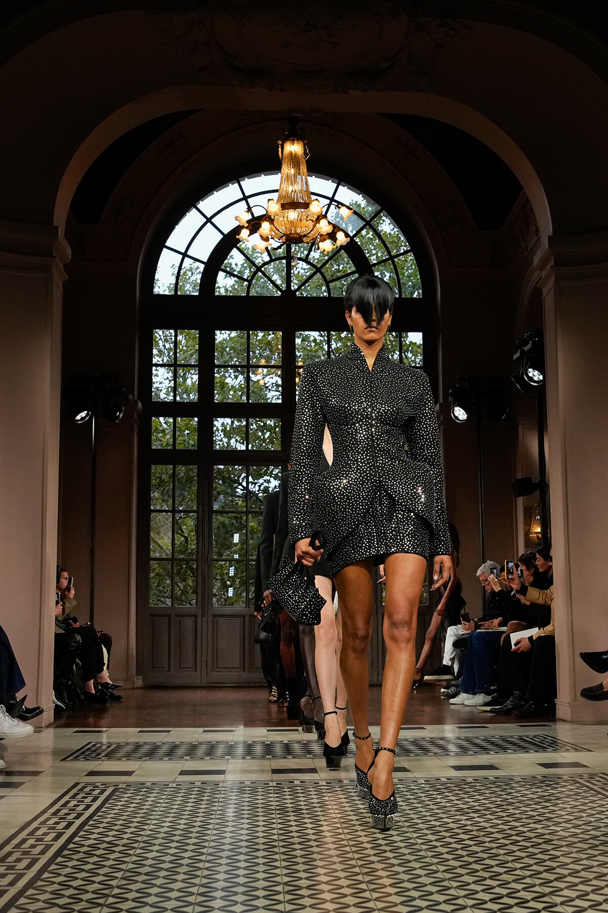 Model walks the runway in a black polka-dot, structured mini dress with a peplum, in an ornate hall with a chandelier above.