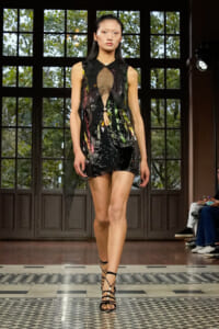Model walking a runway in a black sleeveless dress with colorful abstract print and sheer front panels, paired with strappy heels in a gallery-like venue.