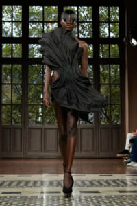 Model walks a runway in a dramatic black, draped avant-garde dress with cutouts and high-contrast silhouette.