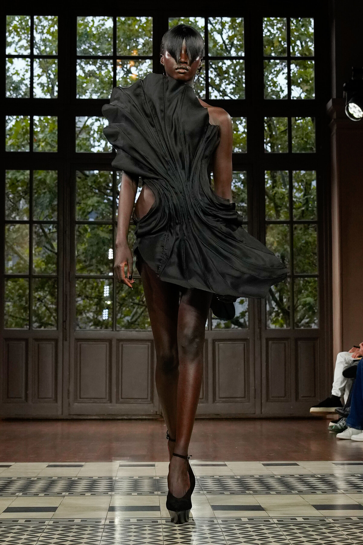Model walks a runway in a dramatic black, draped avant-garde dress with cutouts and high-contrast silhouette.