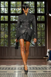 Model walking a runway in a black speckled two‑piece outfit with a structured peplum jacket and short asymmetric skirt, paired with matching dotted heels and a small clutch.