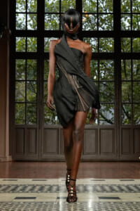 Model walking a runway in a black one-shoulder dress with fringe panels and strappy heels in front of large grid windows.