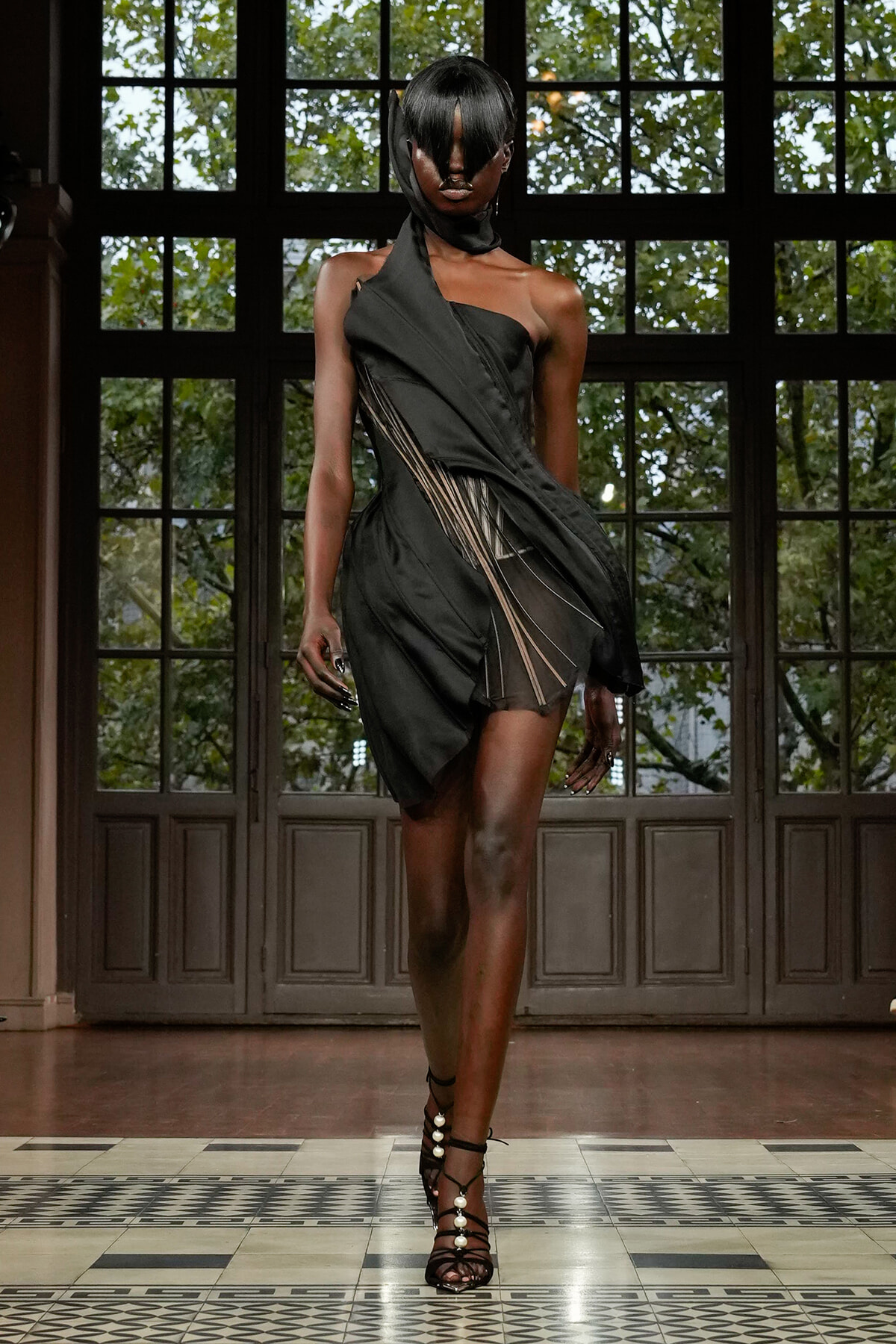 Model walking a runway in a black one-shoulder dress with fringe panels and strappy heels in front of large grid windows.