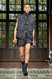 Male model walks a runway in a dark denim layered vest with a high collar, matching shorts, and black platform boots.