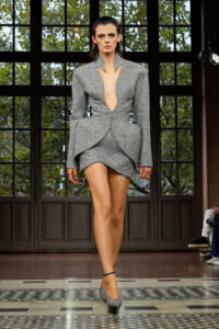 Model on a runway wearing a gray textured peplum blazer with a deep plunging neckline and a matching short skirt.