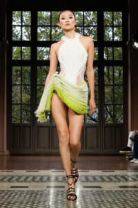 Model walks a fashion runway in a white halter dress with sheer panels and a lime-green ruched skirt, wearing black strappy heels in a paneled room with large windows.