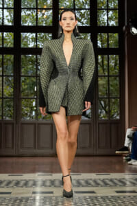 Model on a runway wearing a gray-green, diamond-patterned mini dress with a deep V neckline and black extended cuffs, paired with matching patterned heels in a tiled venue.